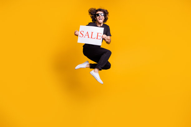 全身大小的人像男子跳高，叫喊保持桌子销售穿着黑色衣服，太阳眼镜孤立在明亮的黄色背景图片下载