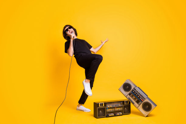 年轻人的全长照片，拿着麦克风，张着嘴，抬起膝盖，两个老式收音机，穿着黑色t恤裤子，白色运动鞋，太阳镜，孤立的黄色背景图片下载