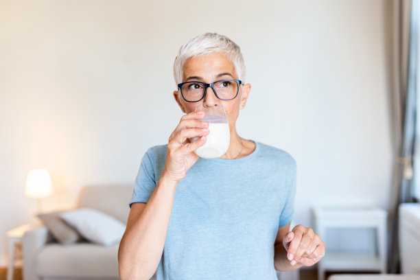在家里拿着牛奶的资深女性。一位中年妇女喝着一杯新鲜的牛奶，开心地站着，微笑着，自信地笑着露出牙齿图片下载