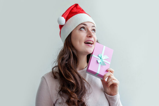 美丽的女孩在红色的圣诞老人帽拿着粉红色的礼盒在手孤立在白色的背景。年轻女子画像，真情流露。圣诞和新年快乐的概念图片下载
