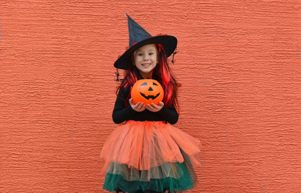 万圣节不给糖就捣蛋。像一个微笑的女孩戴着女巫帽靠在橘子墙。文本复制空间图片下载