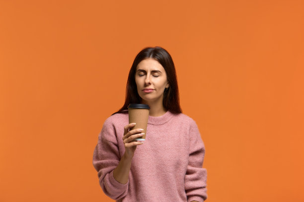 美妙的香气。照片满意的女人在粉红色毛衣拿纸杯热饮，闭上眼睛。美丽的年轻女子喜欢芳香的茶或咖啡。被隔离在橙色墙壁上图片下载