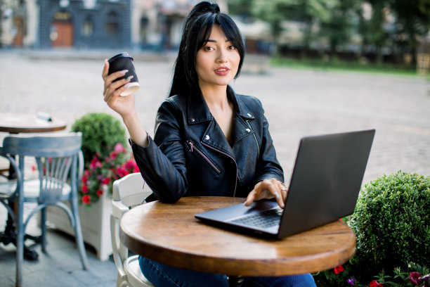 人、城市风格、工作休闲理念。漂亮的亚洲商人穿着黑色皮夹克，使用笔记本电脑，在咖啡店消磨时间时喝咖啡。图片下载