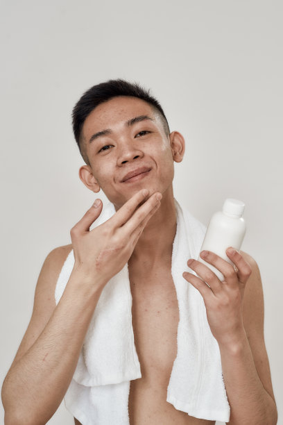画像的赤膊年轻亚洲男子与毛巾在他的脖子上应用乳液剃须后孤立的白色背景。美容、护肤、晨间例行图片下载