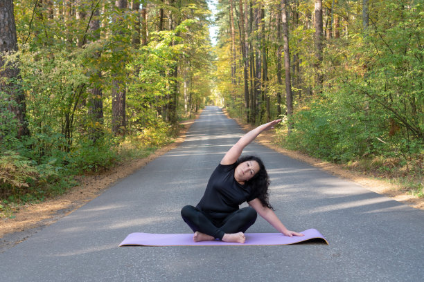 在秋天的森林里，一个穿着黑色t恤和黑色运动裤的胖白种女人正在紫色的垫子上做瑜伽。图片下载