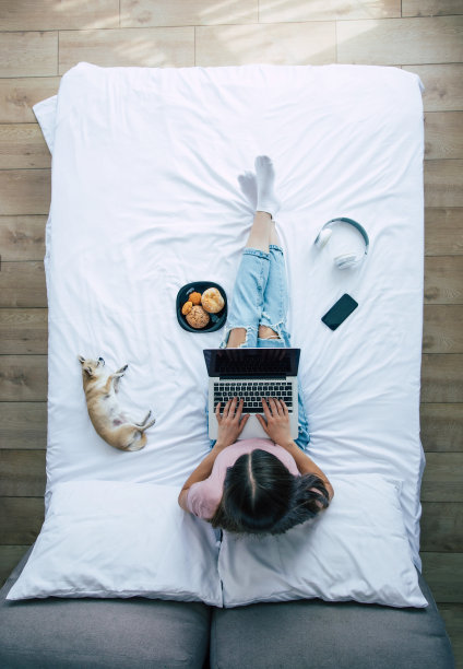 全长度俯视图照片的一个年轻的黑发女人在休闲衣服，而她在床上用笔记本电脑工作。快乐女孩在家和狗一起学习图片下载
