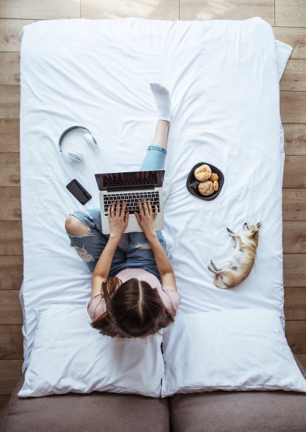 全长度俯视图照片的一个年轻的黑发女人在休闲衣服，而她在床上用笔记本电脑工作。快乐女孩在家和狗一起学习图片下载