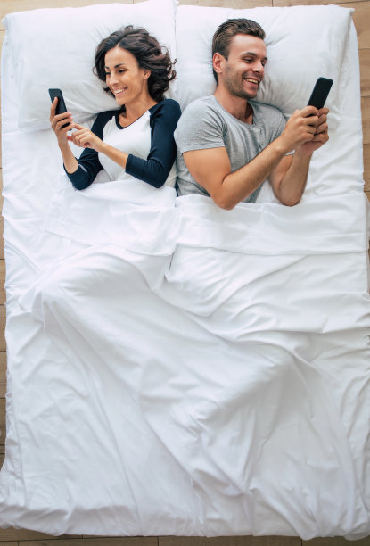 俯视图照片，美女和帅哥躺在白色大床上使用智能手机。网上聊天。上图是这对夫妇的照片。图片下载