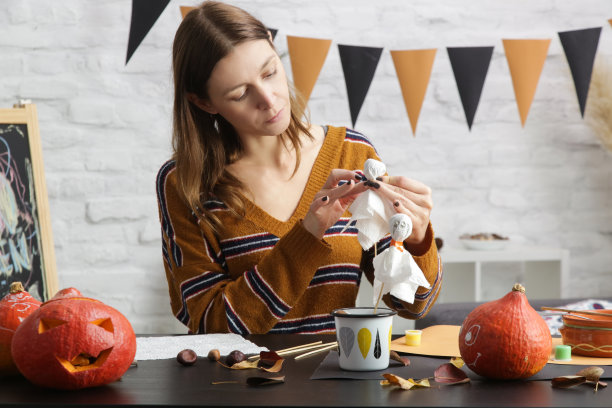 年轻女子在家制作万圣节装饰。秋季DIY工艺。女人在纸棍上做鬼，照片系列。图片下载