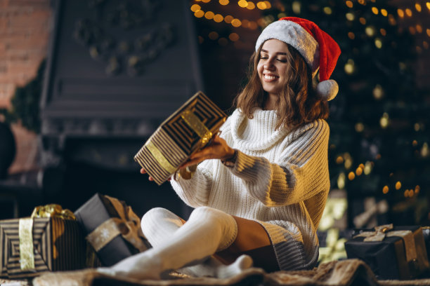 漂亮的女人穿着暖和的毛衣，袜子和圣诞帽，坐在家里的地板上拿着礼品盒图片下载