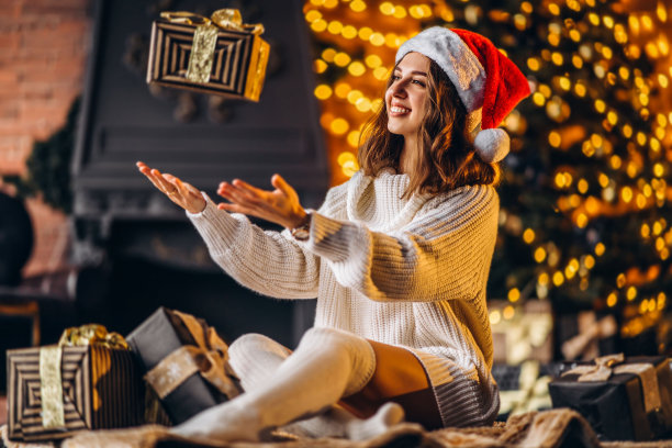 漂亮的女人穿着暖和的毛衣，袜子和圣诞帽，坐在家里的地板上拿着礼品盒图片下载