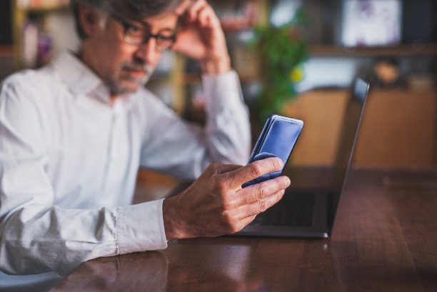 深思熟虑的商人戴着眼镜坐在笔记本电脑前，以一种智能的方式在家工作，使用智能手机连接和发送信息，专注于手和智能手机，模糊的背景图片下载