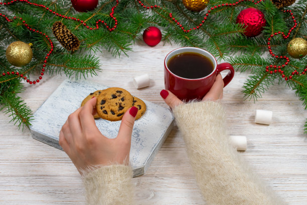 女人手里拿着一杯茶和饼干，放在有圣诞树和装饰品的木桌上图片下载