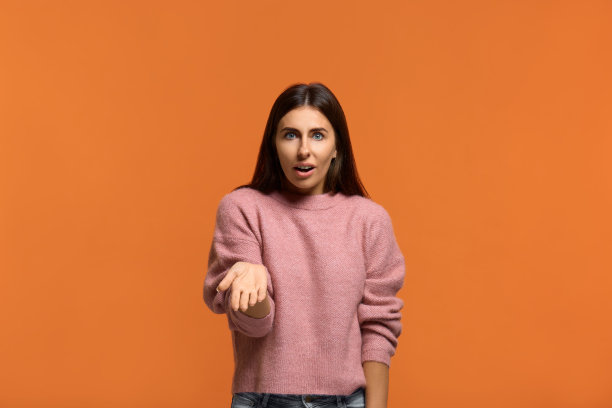 照片中的愤怒的欧洲女人，耸耸肩，表情茫然，模特在白色的背景下，困惑地问你为什么要这样做。穿着粉红色的针织毛衣，隔着橙色的墙图片下载