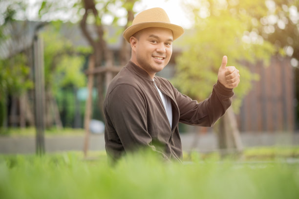 亚洲自由职业者戴上棕色的帽子在户外工作。办公室职员或商人坐在公园区域，用左手竖起大拇指。户外活动的概念。图片下载
