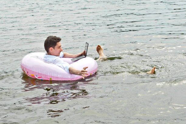 一个穿着衬衫的男人站在一个粉红色的充气圈上。一个拿着笔记本电脑的家伙在河里游泳。假期工作、外包和自由职业的概念。放置文本和复制空间附近的商人。图片下载