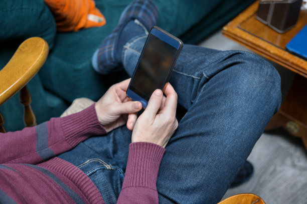 一个男人舒舒服服地坐在家里，用他的智能手机等待消息图片下载