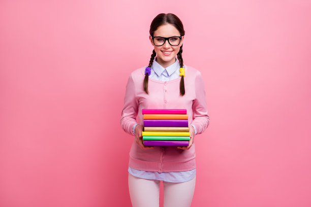 漂亮迷人的学生女士的照片，两个辫子，抱许多书，勤奋的学生参观，参加图书馆，喜欢读书，穿衬衫，套头衫，裤子，specs孤立的，柔和的粉红色的颜色背景图片下载