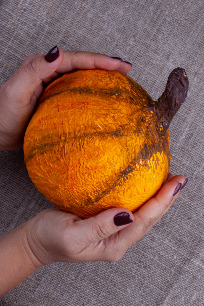 两只修了棕色指甲的手拿着一个自制的橙色纸糊南瓜，背景是粗麻布图片下载