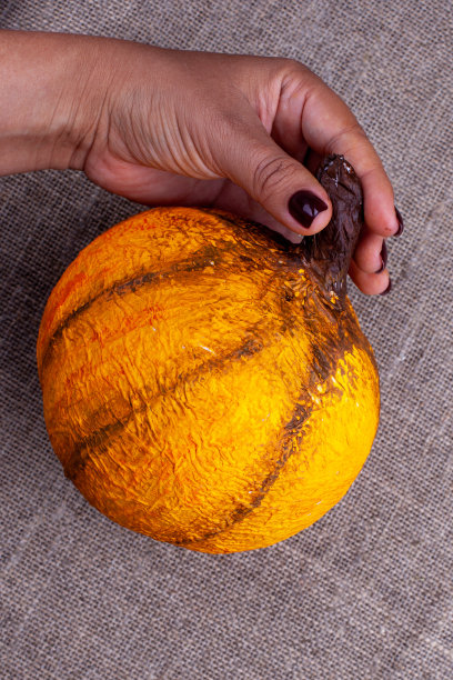一只手拿着一个自制的橙色纸糊南瓜，背景是粗麻布图片下载