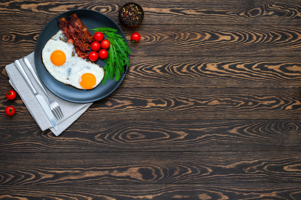 美式早餐，木桌上有煎蛋、香脆培根、圣女果和芝麻菜沙拉，餐巾纸上放着刀叉。图片下载