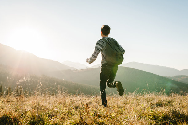 一个年轻的游客背着背包跑在上面欣赏日落。后视图。潮人在山间的秋草中度假。旅行的概念。世界旅游日。图片下载