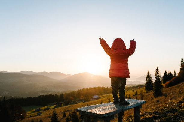 日落时分快乐的小女孩。享受时光的孩子站在日落在雾蒙蒙的山顶。孩子在阳光下旅行在秋天的国家欧洲。后视图。图片下载