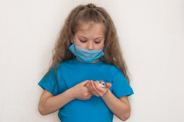 一个戴着面具的学龄前女孩抱着一只白色的准噶尔仓鼠看着他的肖像。工作室白色背景。宠物的啮齿动物。游戏儿童和动物。隔离病毒隔离。图片下载