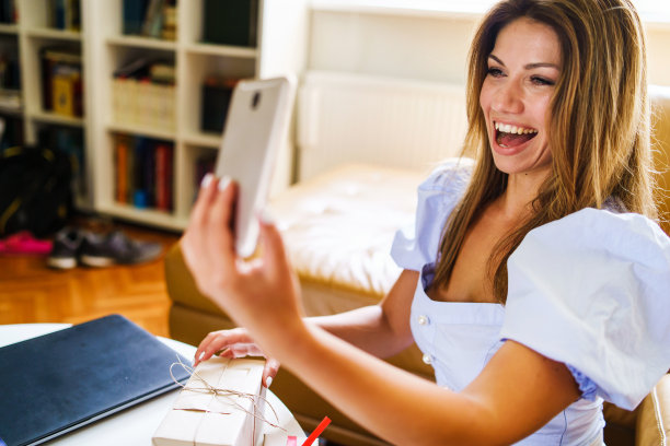 一个快乐的千禧一代白人妇女独自坐在家里打视频电话，微笑着展示礼物包装的礼物-选择性专注于手图片下载