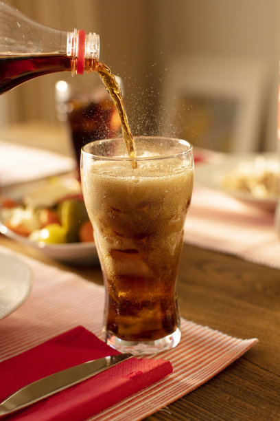 把可乐从瓶子倒进玻璃杯里，在模糊的客厅背景下的桌子上用冰块起泡图片下载