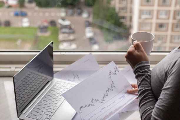 站在公寓窗户旁，用笔记本电脑工作，在纸上打印图形和图表。清晨喝咖啡的女孩。远程工作图片下载