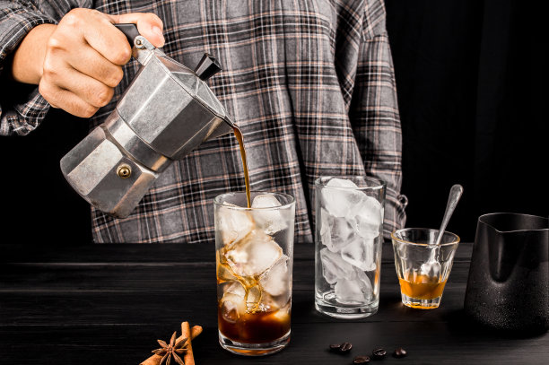 男人用摩卡咖啡壶倒咖啡，煮冷的咖啡上加冰肉桂和蜂蜜在一个杯子里，和水壶牛奶流在木桌上。照片拍摄在工作室的黑色背景。图片下载