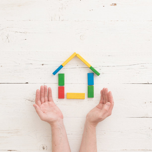 一名男子的双手保护着一座由五颜六色的木块组装而成的房子。家庭、家庭、教育理念。俯视图上的多色砖在木制桌子的背景图片下载