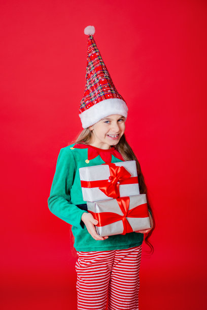 圣诞快乐，一个快乐迷人的女孩，在鲜红的背景下，穿着圣诞老人助手精灵的服装，拿着礼物。图片下载