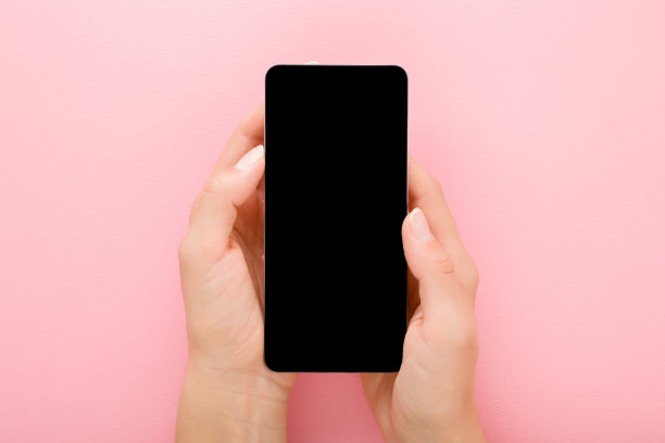 年轻的成年女性手握智能手机，黑色屏幕上的浅粉色桌子背景。柔和的颜色。文本的空位置。特写镜头。视角拍摄。自顶向下的观点。图片下载