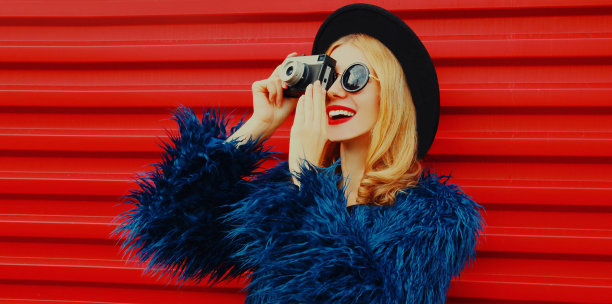 年轻女子的肖像与复古相机拍照穿着蓝色人造皮草大衣，圆帽子在红色的背景图片下载