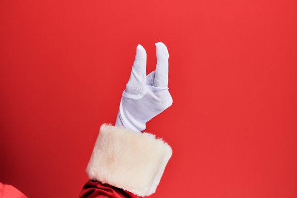 一个穿着圣诞老人服装和手套的人的手在红色背景上采摘和拿走看不见的东西，用手指抓住物体显示空间图片下载