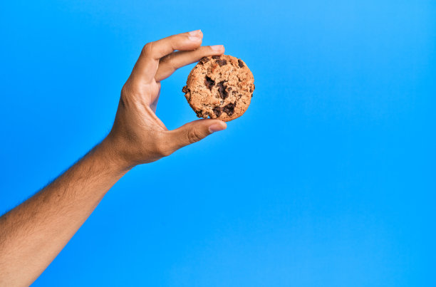 年轻的西班牙裔男子的手拿着巧克力饼干在孤立的蓝色背景。图片下载