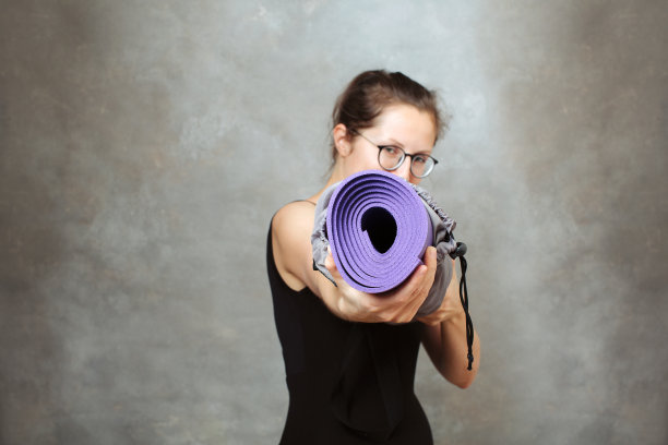 女子瑜伽在玩的时候，用两只手拿着一个垫子对着镜头。以灰色的灰泥墙为背景图片下载
