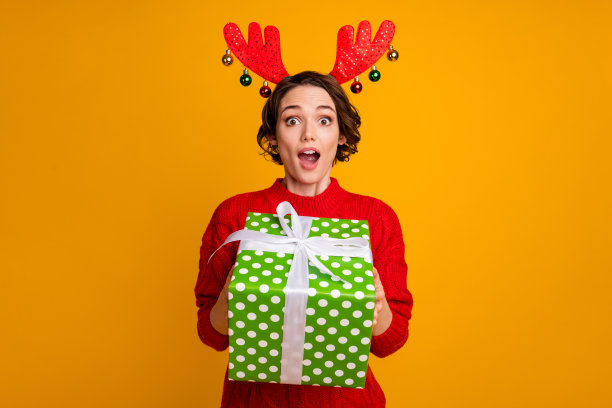 照片兴奋美丽的女士冬季假期概念举行大礼盒节日活动派对圣诞快递送穿鹿角装饰红色针织毛衣孤立的黄色背景图片下载