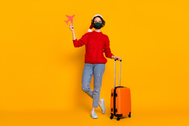 美丽的女士的全身照片来冬季假期飞回家家庭持有滚动的情况纸飞机穿医疗安全面具圣诞帽毛衣牛仔裤鞋子孤立的黄色背景图片下载