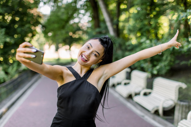 户外肖像的年轻美丽的亚洲女孩穿着黑色连衣裙迷人的微笑，自然的化妆和黑色的马尾辫，享受她在公园散步，玩她的手机，自拍照片图片下载