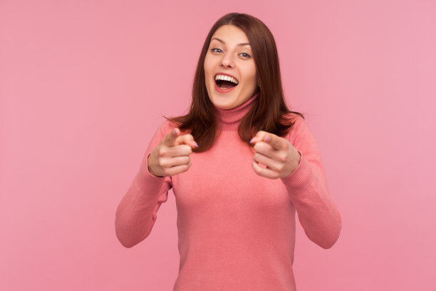 开朗积极的黑发女人在粉红色毛衣指着手指，微笑着看着相机，嘲笑和嘲笑图片下载