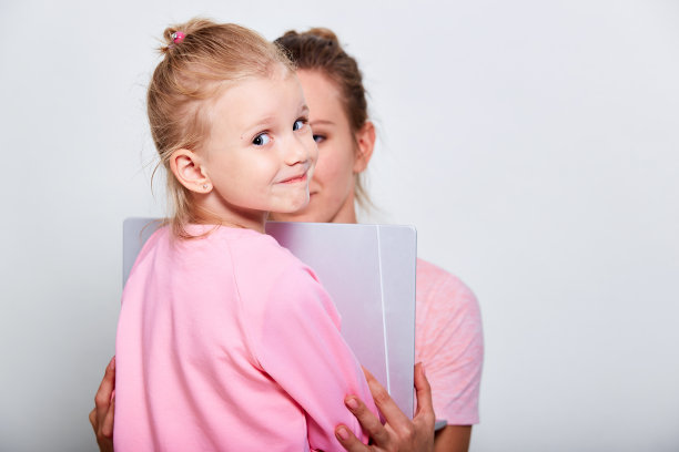 病毒防护概念，4-5岁可爱女孩和她穿着粉色衣服的母亲的特写肖像，展示屏幕上有医用口罩的笔记本电脑，灰色背景图片下载