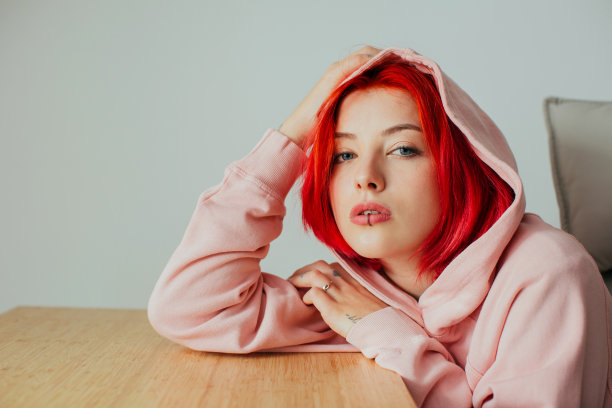 一个酷的十几岁女孩的肖像与明亮的红色头发，嘴唇穿孔和帽衫抱着头在桌子上看着相机图片下载