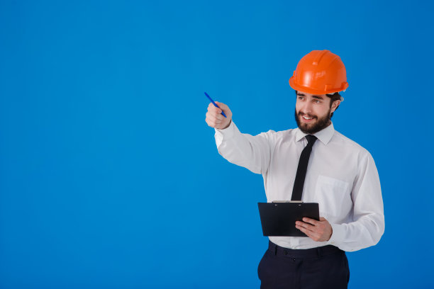 一名留着胡子的年轻男性建筑工人，身穿白色衬衫，戴着建筑头盔，背景是蓝色。工业工程师-建筑师在一个孤立的背景上拿着带有文件的剪贴板图片下载