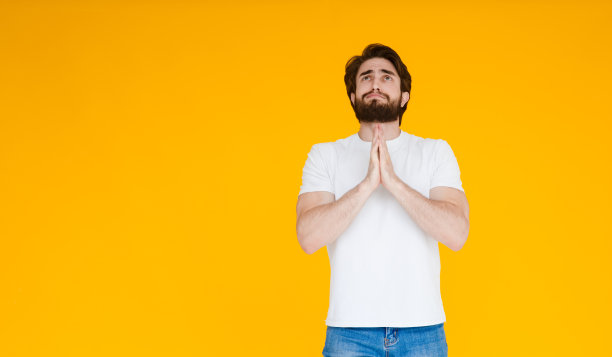 恳求年轻的大胡子男人在白色休闲t恤摆孤立的黄色墙壁背景工作室肖像。人的情感生活理念。模拟拷贝空间。双手合十祈祷。图片下载