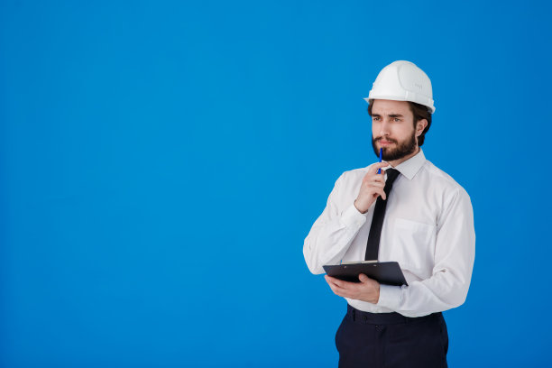 一名留着胡子的年轻男性建筑工人，身穿白色衬衫，戴着建筑头盔，背景是蓝色。工业工程师-建筑师在一个孤立的背景上拿着带有文件的剪贴板图片下载
