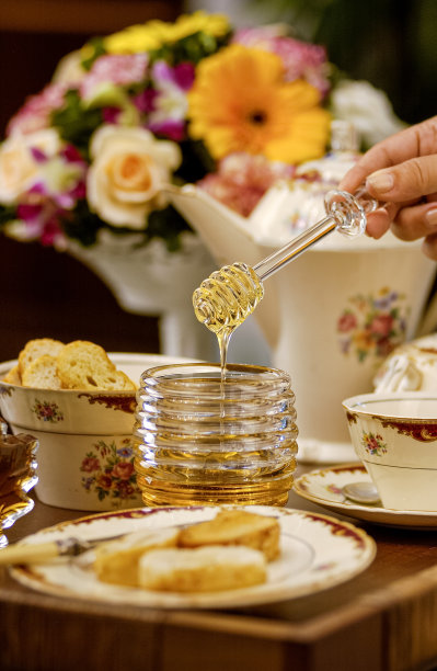 玻璃容器里装着蜂蜜，双手捧着蜜露，把蜂蜜放在烤面包上，桌子上摆着复古风格的盘子和以模糊鲜花为背景的水壶。图片下载