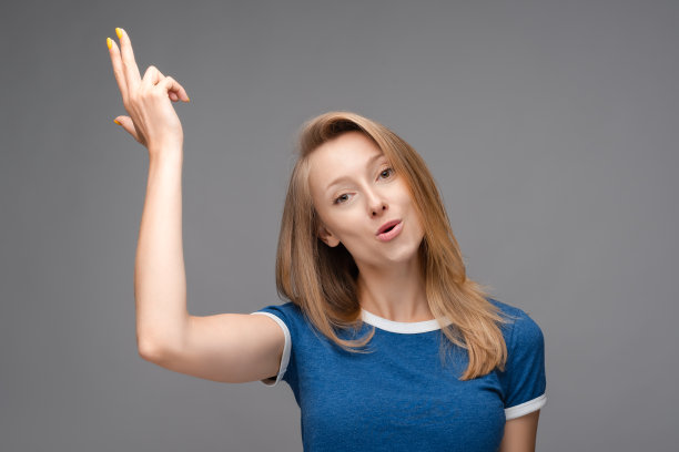 困惑的金发女孩拿着象征性的枪与手势。工作室拍摄，灰色背景。人类情感的概念图片下载